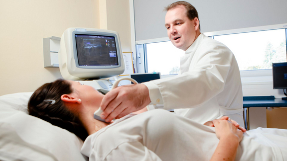 Arzt führt eine Ultraschalluntersuchung am Hals einer liegenden Patientin durch und erklärt den Befund am Monitor.